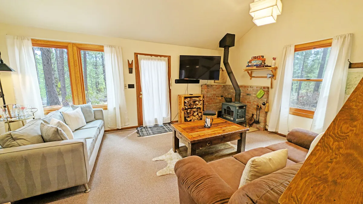 The living room features a wood-burning stove, coffee table, and large windows that let in natural light and showcase woodland views. Plush seating offers a comfortable spot to relax, socialize, or enjoy the scenery, embodying the cabin's cozy core.