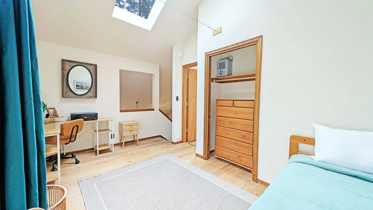 A tranquil upstairs bedroom with a bed featuring light blue bedding, a wooden dresser, a desk with a chair for work or study, and a window letting in ample daylight. The space balances comfort and productivity.