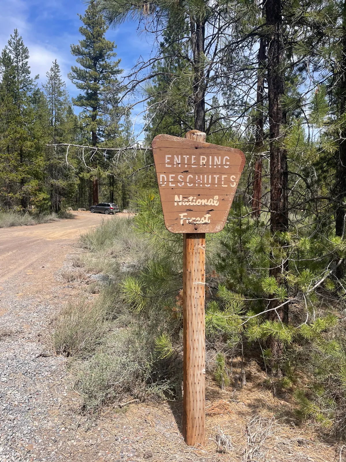 A rustic wooden sign marks the entrance to a gravel trail surrounded by dense forest. The setting feels adventurous yet serene, inviting exploration and reinforcing the idea of stepping directly into nature from your front door.
