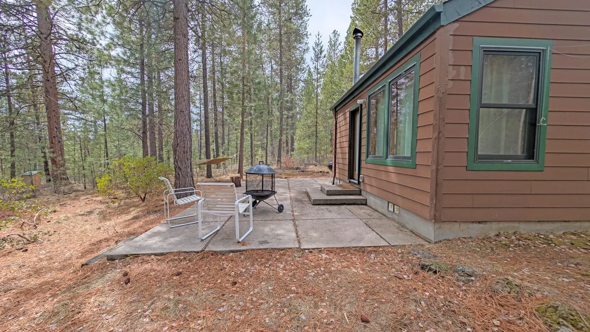A wooden cabin is nestled among trees, with a patio area and grill in the foreground. The outdoor setting highlights the peacefulness of the place, while the patio space suggests opportunities for outdoor relaxation and activities with your dog.