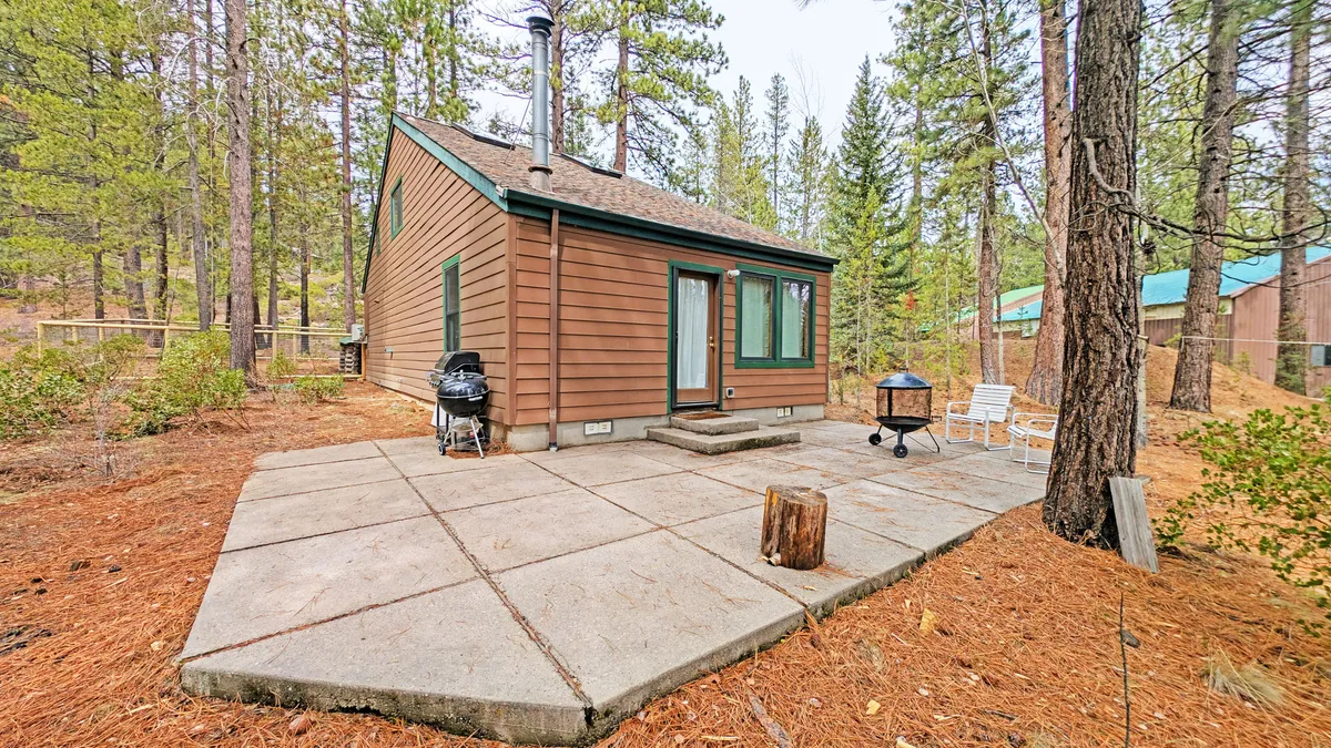 This exterior image features a wooden cabin with a prominent grill set up on a spacious paver patio. Comfortable outdoor seating is surrounded by mature trees, creating a private and inviting outdoor living area. The scene is set for barbecues, outdoor relaxation, and gatherings around the firepit under the stars.