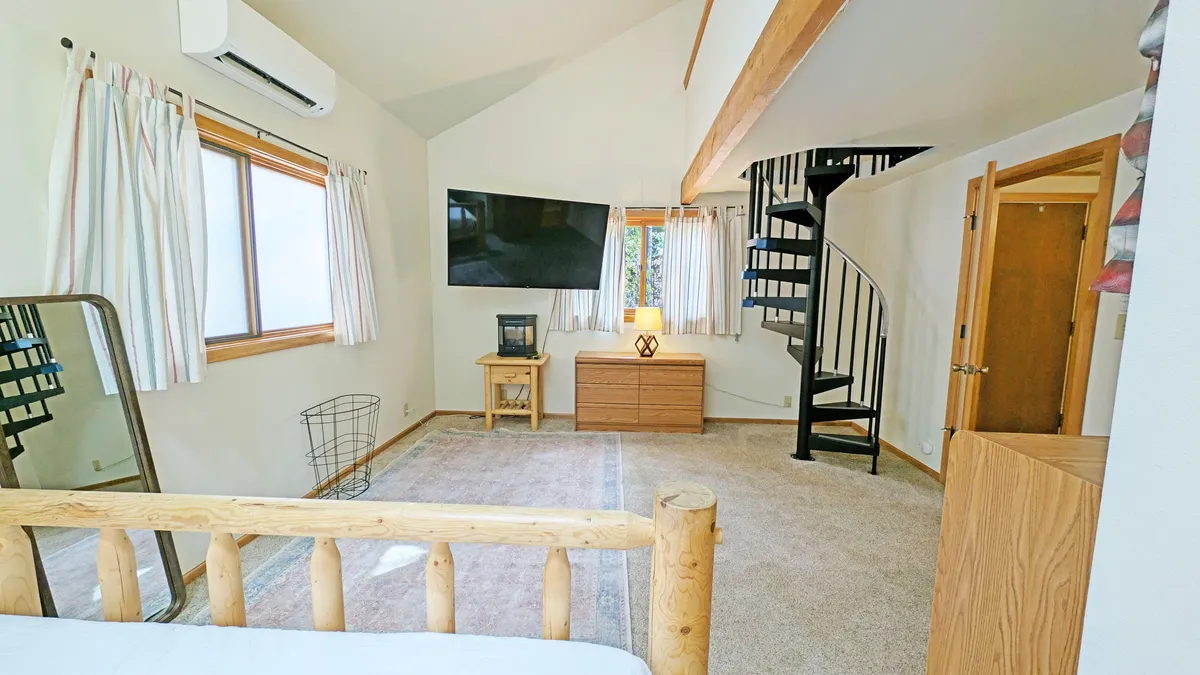 A bright and spacious room featuring a wooden log bed, a spiral staircase, a large window streaming in natural light, and a TV mounted on the wall. The space exudes comfort and showcases the multi-level design central to the cabin’s layout.