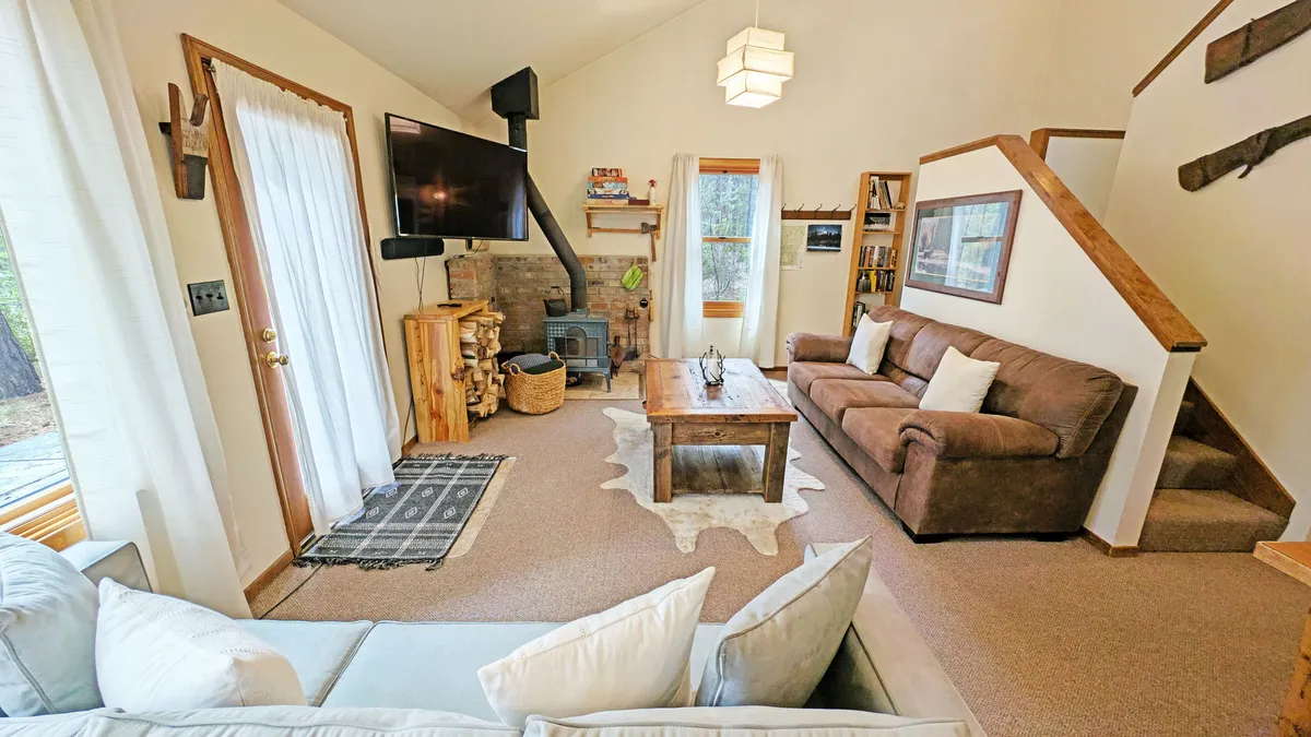 The image showcases the inviting main living area of the cabin, featuring plush sofas gathered around a rustic wooden coffee table, a glowing fireplace, and a TV—set beneath a cozy loft and next to a wooden staircase. The ambiance is warm and welcoming, perfect for relaxation and gathering with loved ones.