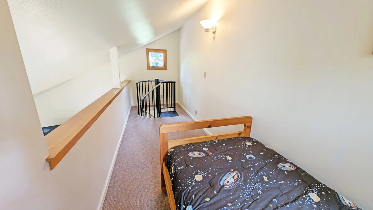 A playful loft area with a twin bed dressed in space-themed bedding, a window letting in soft natural light, and a staircase leading down. The space feels cozy and perfect for a child or solo guest.